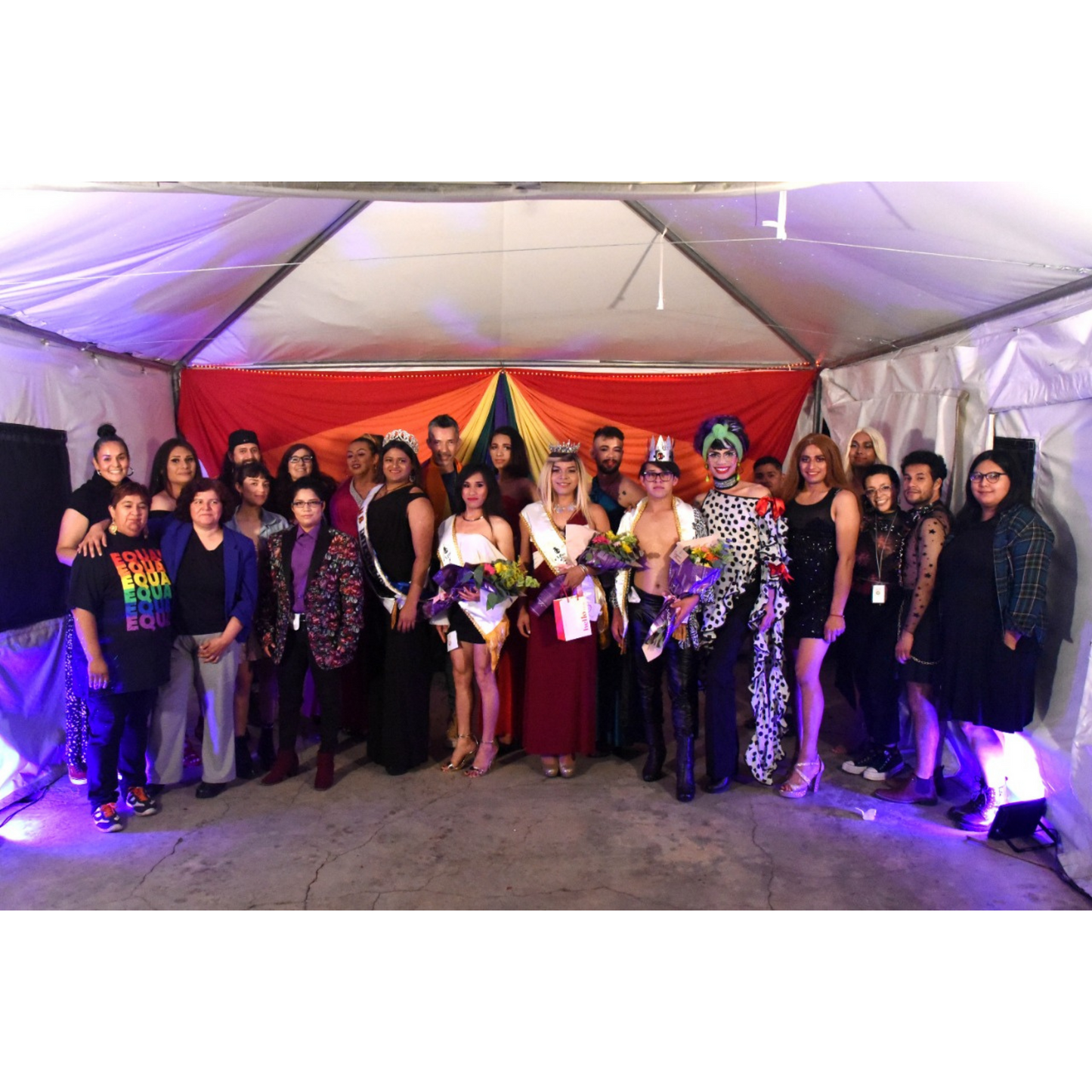 A group of 25 people from the LBTQIA+ community standing in a large tent. Some of them are wearing ball clothing with ribbons attached, and holding large flower bouquets.