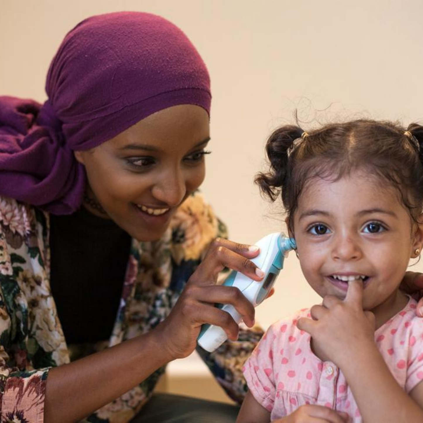 A smiling young girl getting her in ear tempterature check by a woman wearing a headscarf. They are both smiling and the girl has her hair tied up in two ponytails.