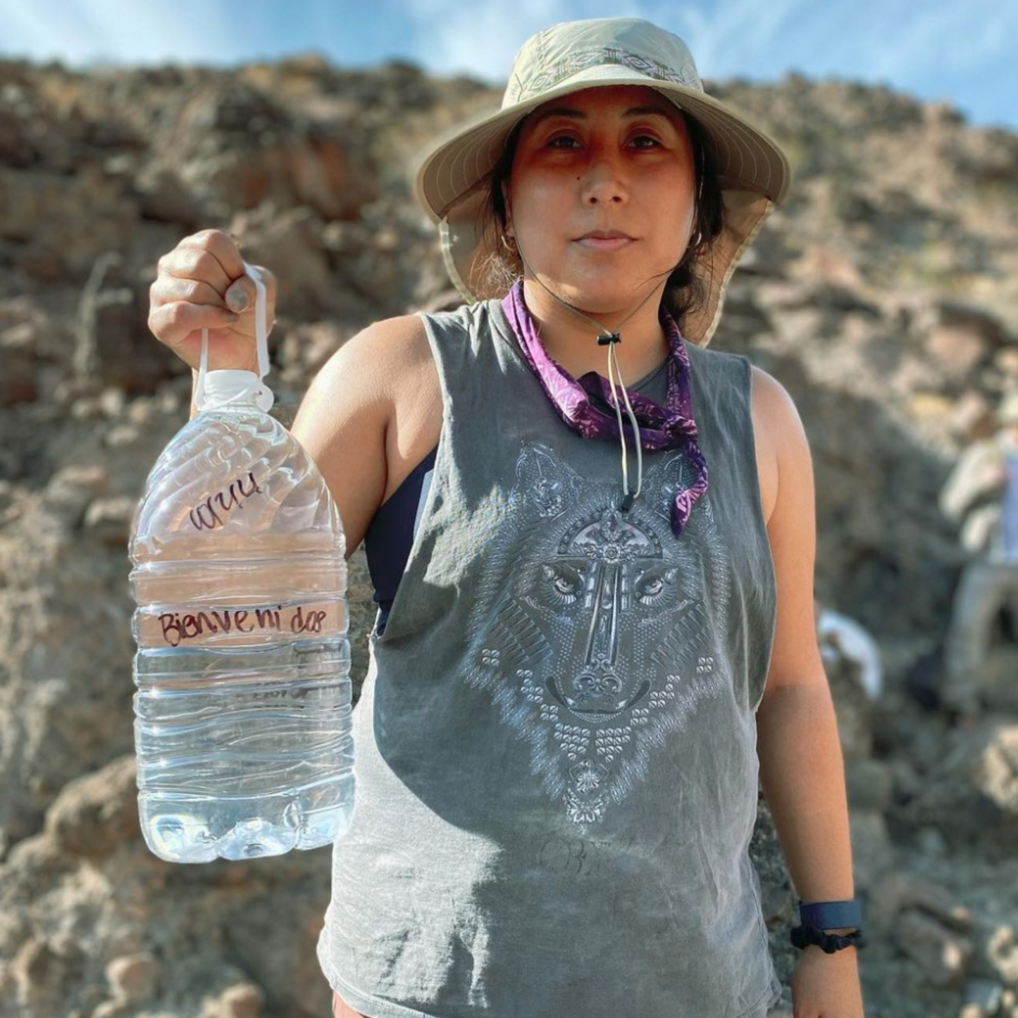 A woman with her hair tied back wearing a boonie hat. She is holding a five litre water bottle with the text Bienvenidos written across which means Welcome in English.