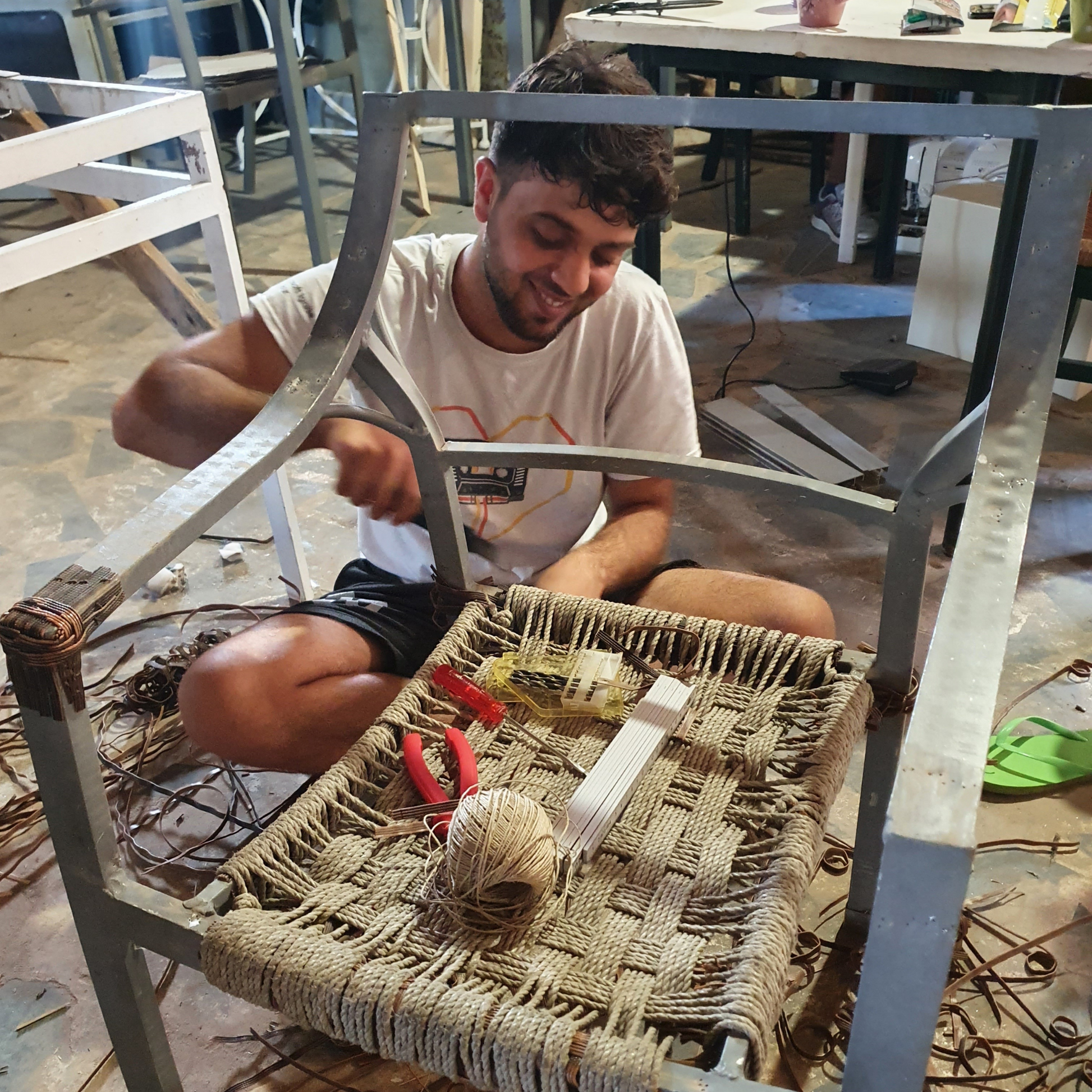 This photography was taken in a building and repair garage, showing a smiling refugee with wavy hair and a beard repairing a wooden chair that has a rope seat. In the background there are partial views of a desk with tools on it, a wooden bed and a wooden shelf. On the floor there are wooden scraps from the work done.