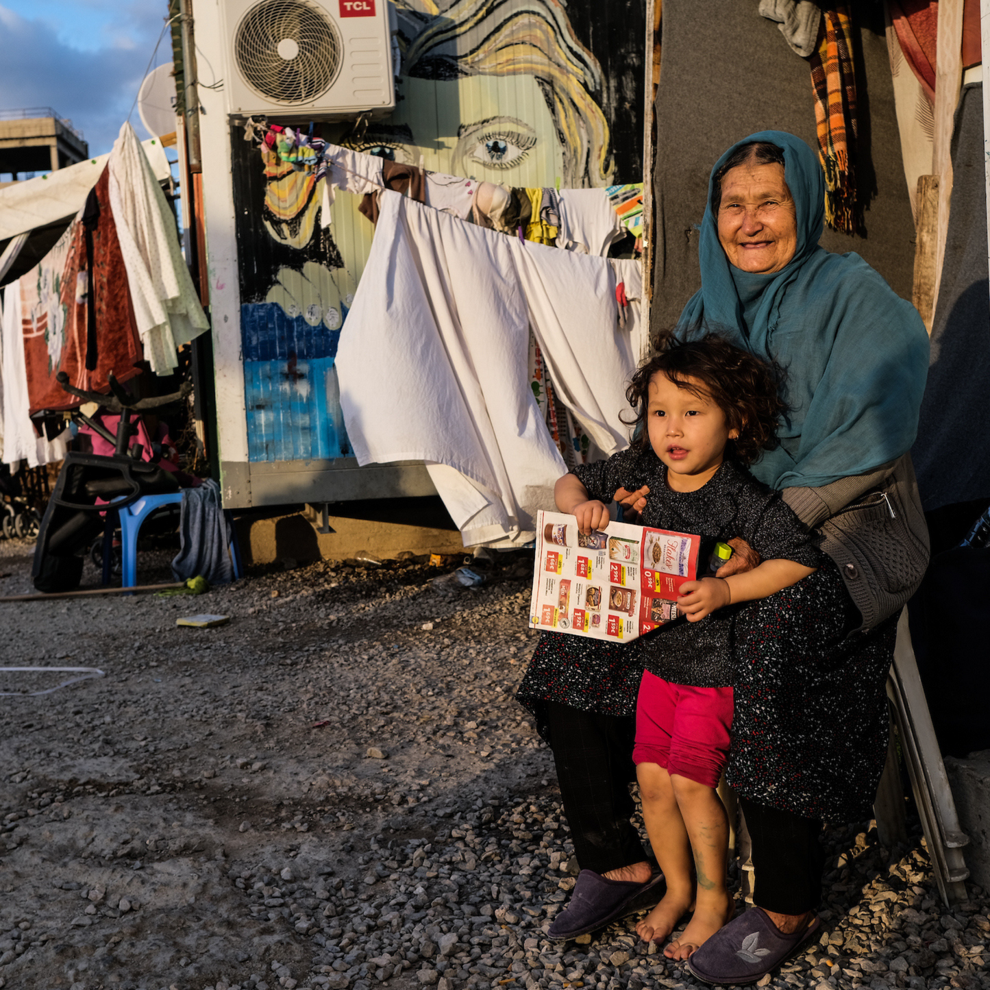 This photography taken in a refugee camp shows an older woman sitting down whilst holding a child. In the background there are clothes, bedding and thick blankets hanging off ropes to dry.