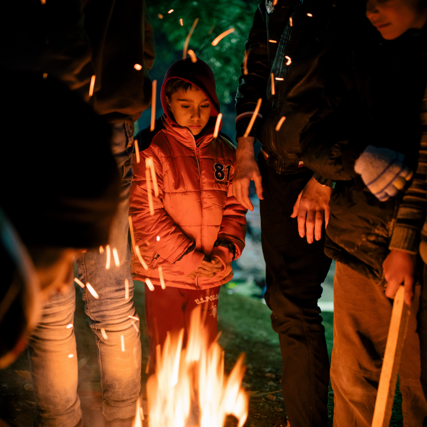 The image is showing a couple of refugees around a fire at night trying to warm up. The focus is on a young boy who is wearing a thick winter coat and is looking down at the fire.
