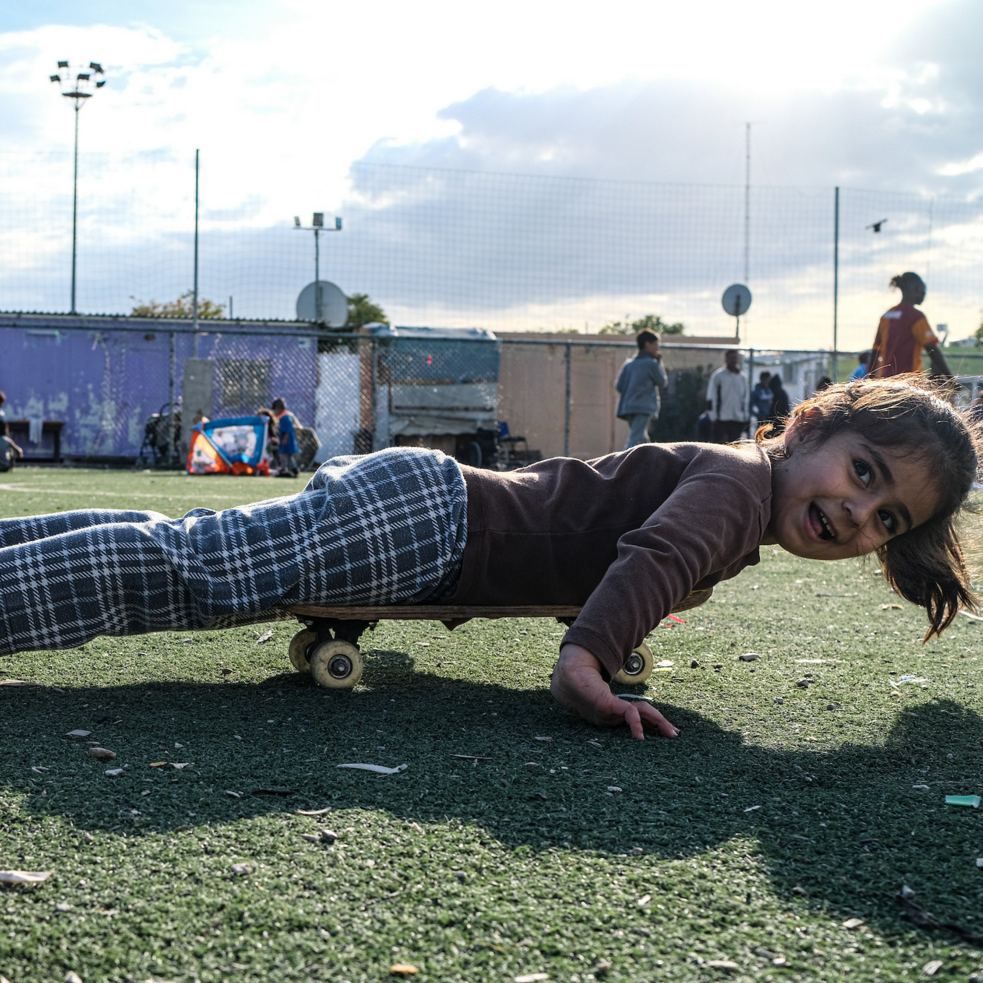 The image shows a young girl being playing and laying on a child's skating board, in a playing space of a refugee camp. In the background there are makeshift accommodations and a few other adult refugees.