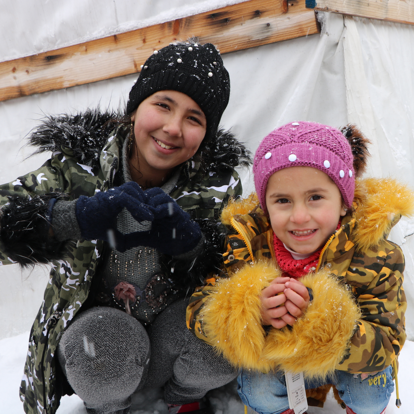 This image has 2 young refugee girls dressed for winter with their hoodies on, smiling and symbolising hearts wirth their hands placed across their chest. They are both wearing winter hats and are in front of makeshift accomodation with plastic wrap on it.