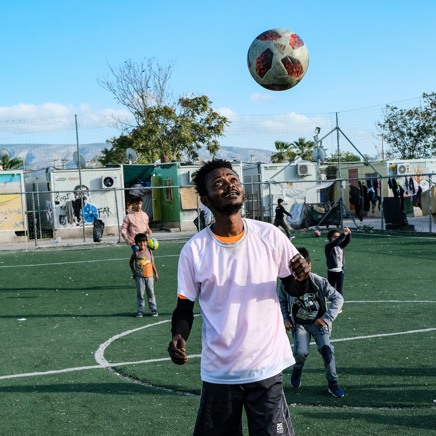An image with a refugee man playing with a football on a pitch. Behind him there are 4 boys looking at him. In the background there is a refugee camp with improvised accommodation.