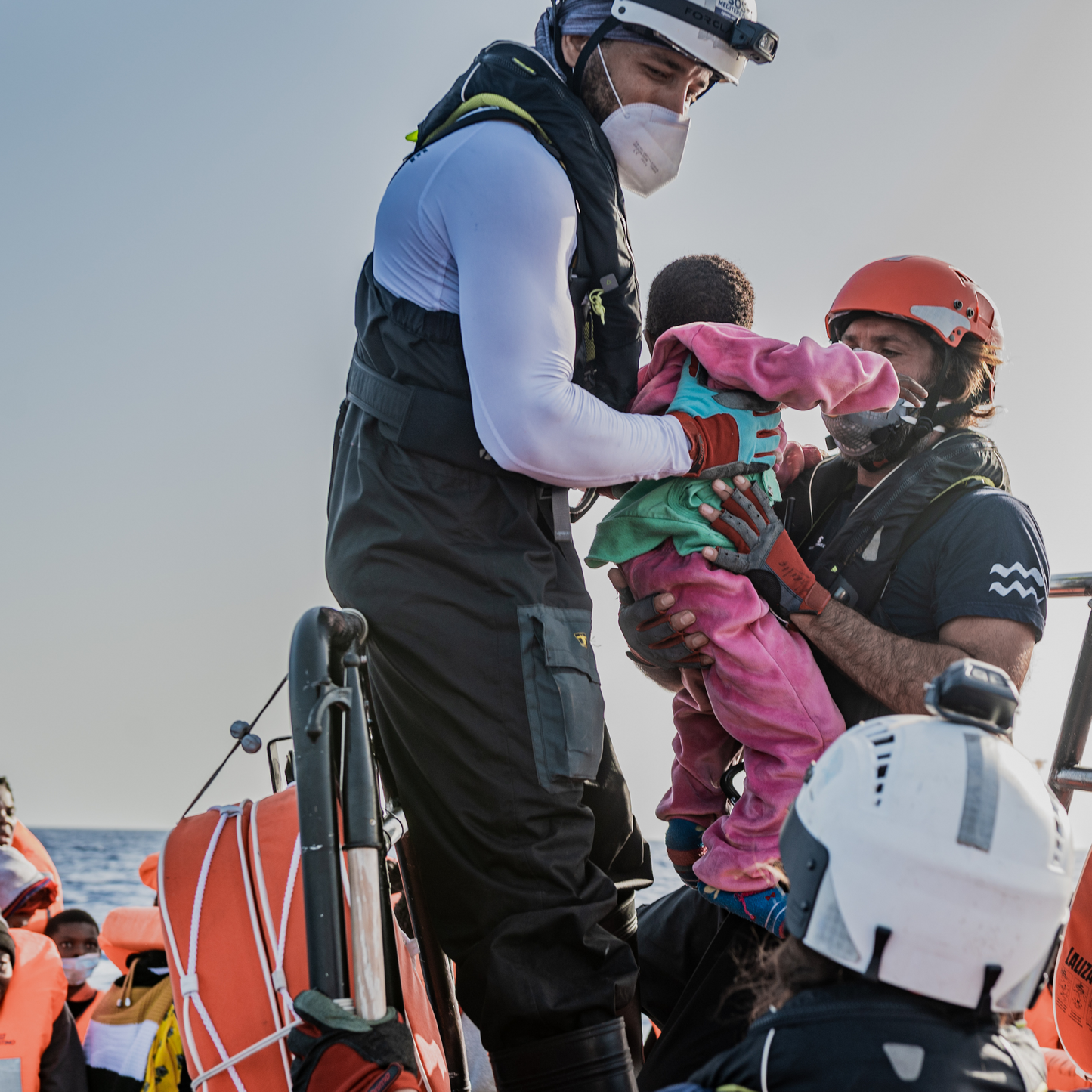 Refugee toddler beign saved from the sea. Other refugees can be seen in the background on boats.