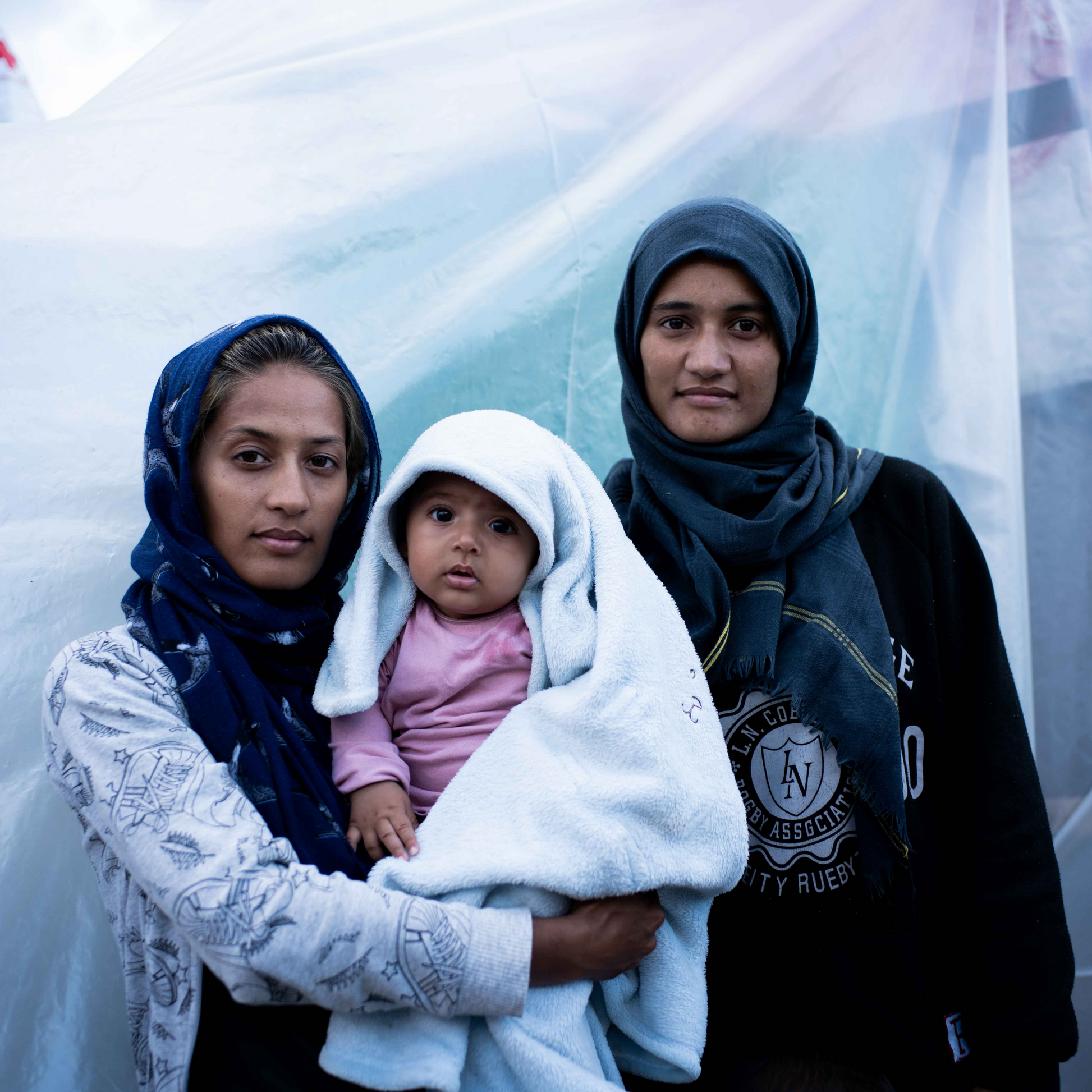 This photography was taken in a camp. In the image there are two adult refugee women with their heads covered and the one of the left is holding a baby who wears a pink top and covered with a thick white blanket. They are pictured in front of a make shift accommodation that is covered with a thick white plastic sheet.