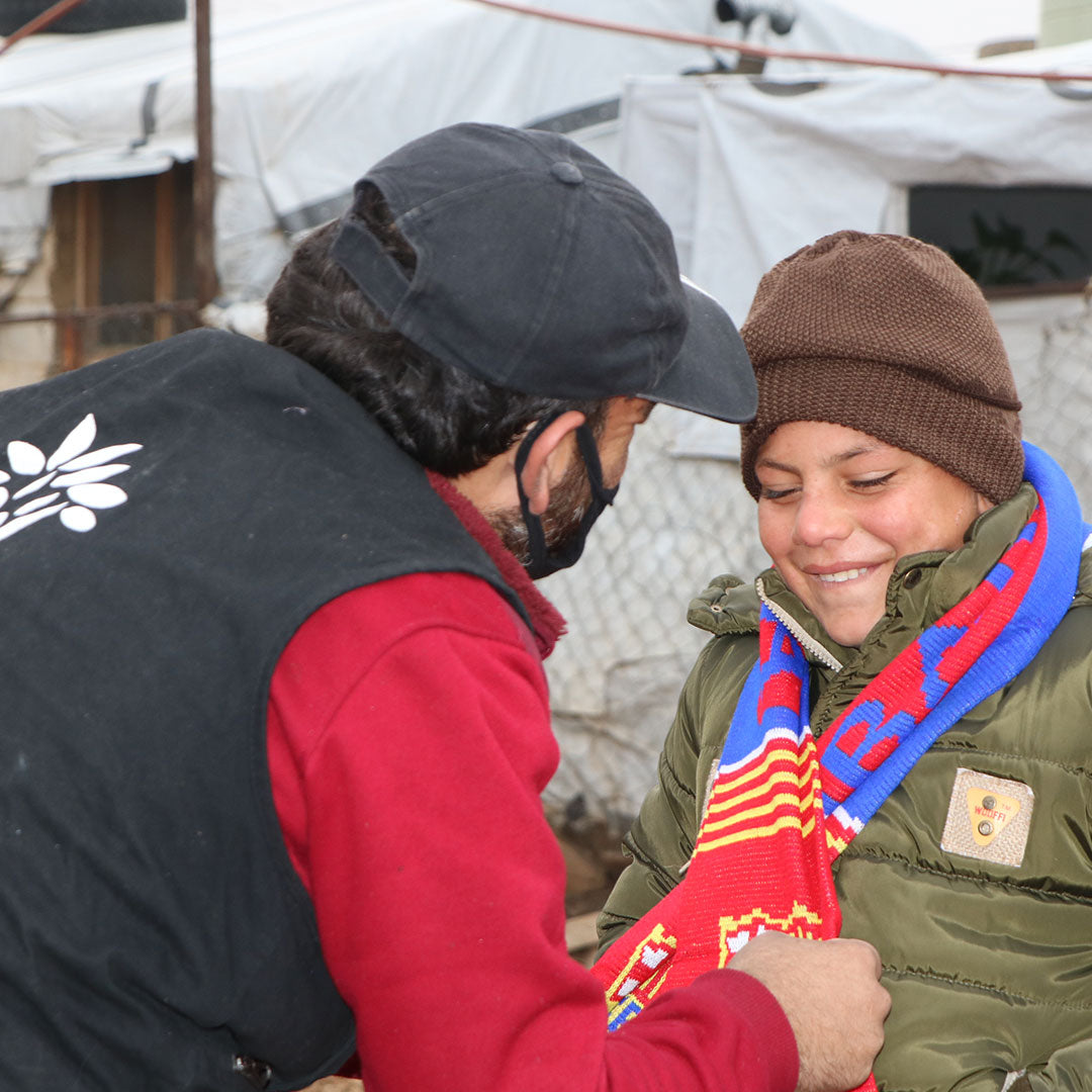 An adult man and a young boy dressed in winter clothes, in a refugee camp. The man is making the boy comfortable by putting a scarf around his neck. In the background there is imporvised accommodation for refugees, covered by plastic.